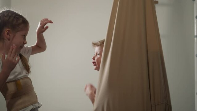 Playful toddler boy attacks sister sitting in hanging hammock in home gym slow motion. Girl wearing posture corrector has fun with little brother