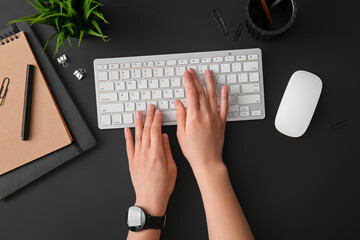 Female programmer using computer keyboard with mouse, notebooks and stationery on dark background