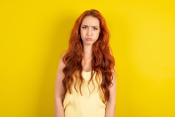 Offended dissatisfied  young redhead woman wearing  yellow t-shirt over yellow wall with moody displeased expression at camera being disappointed by something