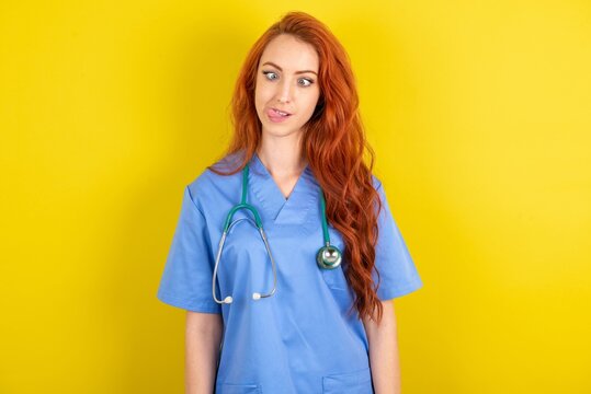 Funny redhead doctors woman wearing blue uniform over yellow wall makes grimace and crosses eyes plays fool has fun alone sticks out tongue.