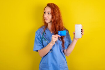 redhead doctors woman wearing blue uniform over yellow wall holding bank card modern device looking empty space