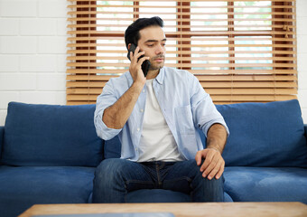 Fototapeta premium young handsome man sitting on sofa and talking by smartphone at home