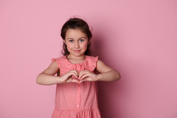 Adorable beautiful Caucasian blue eyed little child girl in pink dress, forms a heart shape with her hands, smiling looking at camera, expressing positive emotions, isolated pink background Copy space