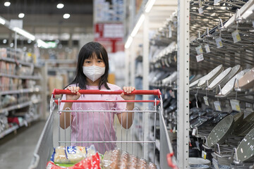 Asian child or kid girl wearing n95 white face mask to closed mouth nose for protect pm 2.5 dust by air pollution to covid-19 with pushing supermarket trolley or cart for shopping in department store