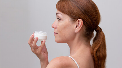 Back of cropped caucasian middle aged woman face with freckles holding opened jar with skin cream in hand on white background