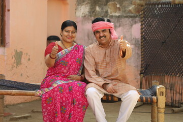 Happy Indian village family of husband and wife showing thumbs up in agreement sitting together outside the house.