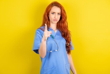 young red-haired doctor woman over yellow studio background frustrated and pointing to the front