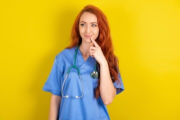 Lovely dreamy young red-haired doctor woman over yellow studio background keeps finger near lips looks aside copy space.