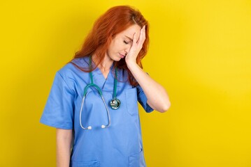 young red-haired doctor woman over yellow studio background with sad expression covering face with...
