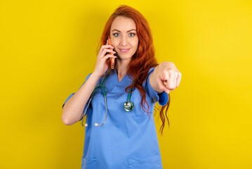Positive young red-haired doctor woman over yellow studio background indicates directly at camera...
