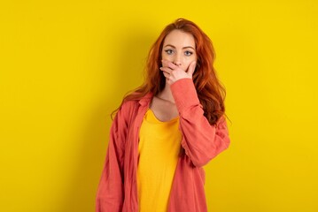Emotional young beautiful red haired woman over yellow studio background gasps from astonishment,...