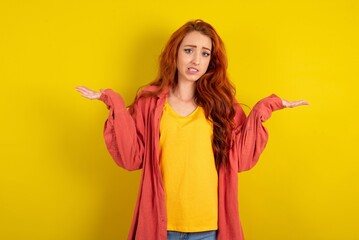 Clueless young beautiful red haired woman over yellow studio background shrugs shoulders with...