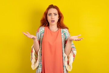 Frustrated young beautiful red haired woman feels puzzled and hesitant, shrugs shoulders in...