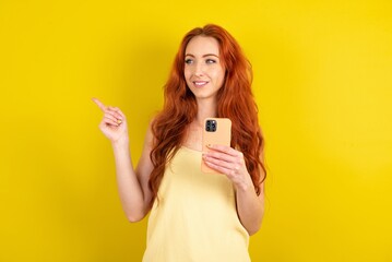 Smiling young beautiful red haired woman wearing yellow blouse over yellow studio background holding mobile phone and pointing aside at copyspace.