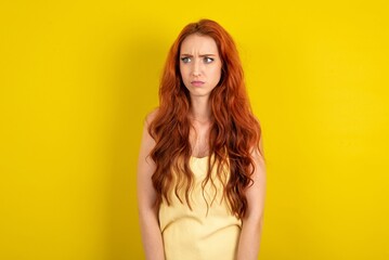 Fototapeta premium young beautiful red haired woman wearing yellow blouse over yellow studio background stares aside with wondered expression has speechless expression. Embarrassed model looks in surprise