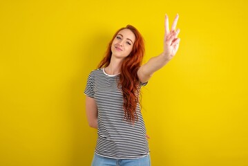 young beautiful red haired woman wearing striped t shirt over yellow studio background directs...