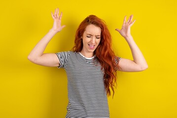 young beautiful red haired woman wearing striped t shirt over yellow studio background goes crazy as head goes around feels stressed because of horrible situation