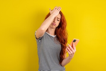 Upset young beautiful red haired woman wearing striped t shirt over yellow studio background makes...