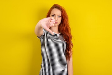 young beautiful red haired woman wearing striped t shirt over yellow studio background looking unhappy and angry showing rejection and negative with thumbs down gesture. Bad expression.