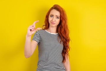 Displeased young beautiful red haired woman wearing striped t shirt over yellow studio background shapes little hand sign demonstrates something not very big. Body language concept.