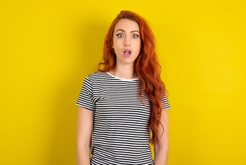 Shocked young beautiful red haired woman wearing striped t shirt over yellow studio background stares bugged eyes keeps mouth opened has surprised expression. Omg concept