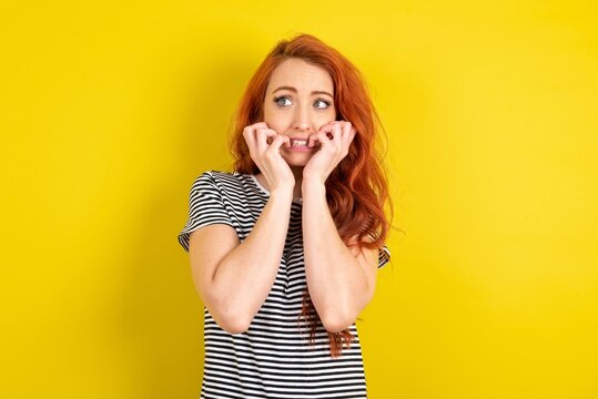 Terrified young beautiful red haired woman wearing striped shirt over yellow studio background looks empty space home alone moonless night