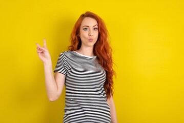 young beautiful red haired woman wearing striped shirt over yellow studio background makes peace gesture keeps lips folded shows v sign. Body language concept