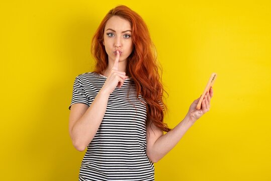 young beautiful red haired woman wearing striped shirt over yellow studio background holding modern gadget ask not tell secrets