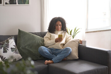 Relaxed young latin woman sitting on sofa using cell phone at home holding smartphone, looking at...