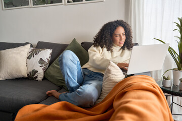 Latin young woman sitting on couch using laptop at home surfing, doing ecommerce shopping, looking...