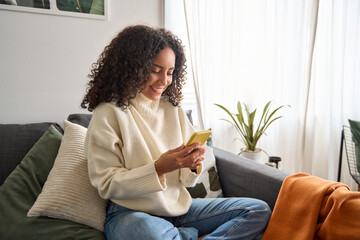 Happy young latin woman sitting on sofa holding mobile phone using cellphone technology doing ecommerce shopping, buying online, texting messages relaxing on couch in cozy living room at home. - obrazy, fototapety, plakaty
