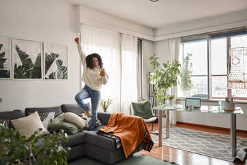 Happy excited young woman using smartphone dancing at home. Latin girl jumping on couch listening...