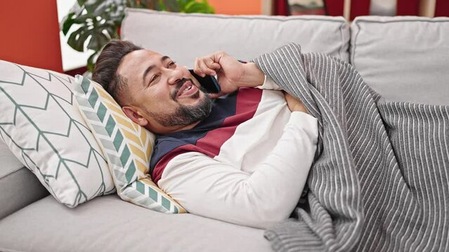 Young latin man talking on smartphone lying on sofa at home