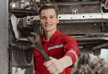 Auto mechanic in red uniform standing at car repair station holding wrench prepare to fix broken engine vehicles at mechanics garage. Smiling maintenance engineer happy work in automotive industry