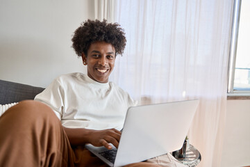 Happy smiling African American gen z teen student sitting at couch relaxing at home with laptop computer looking at camera advertising online learning, remote web elearning. Authentic portrait.