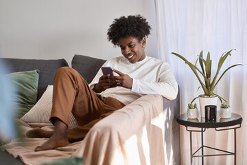 Happy gen z African American teen sitting on couch holding smartphone, using cell phone, looking at...