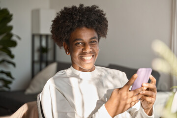 Happy gen z African American teen sitting at home holding smartphone, using cell phone, looking at camera enjoying doing online shopping in mobile apps, influencing or playing game, portrait. - obrazy, fototapety, plakaty