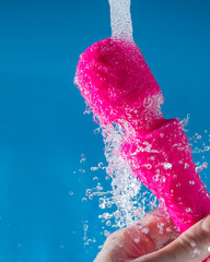 A woman washes a pink vibrator under running water on a blue background. Sex toy hygiene concept.