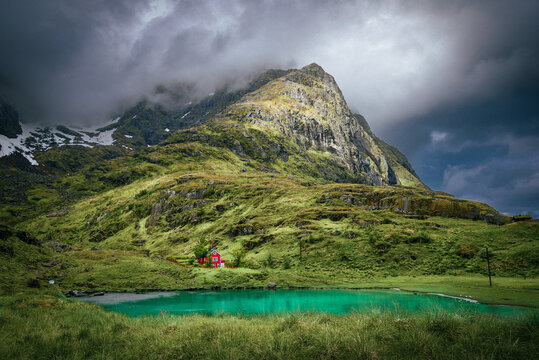 A beautiful view of a mountain and a cottage under it with a scenic and calm lake infront of it in the summer and cloudy skies with sunrays falling on one side of the mountain. Goats are grazing also.