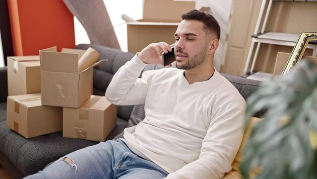 Young hispanic man talking on smartphone sitting on sofa at new home