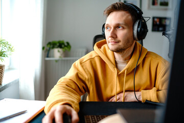Smart bearded Caucasian young man freelancer in yellow sweater and headphones thinking over data while sitting at desk and using laptop in light workplace at home. Copy space