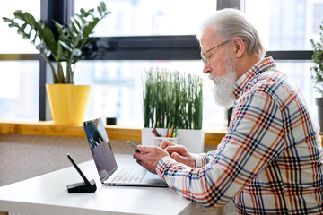 Handsome senior caucasian man in eyeglasses using mobile phone while sitting at cozy workplace with laptop at home, retired male chatting with friends in social media, typing on smartphone.