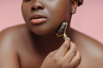 Closeup of beautiful african american woman doing face roller massage isolated on pink background