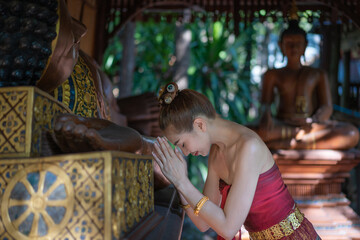 Pretty woman wearing a traditional Thai dress Lanna style clothes pray homage to the reclining Buddha statue carved from golden teak wood for worship and good luck in life.