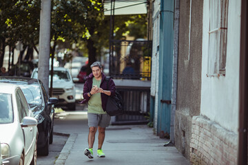 Middle aged man walking home from the gym on the sidewalk of a city street while using a smart phone