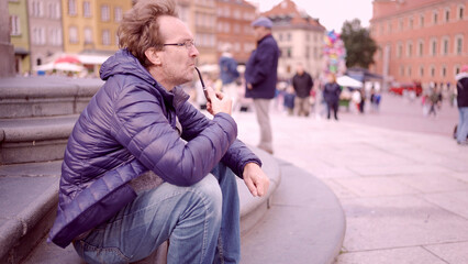 Adult man sitting on square and smoking a tobacco pipe in the Palace Square, Warsaw Old Town