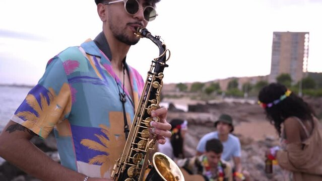Close up of an unrecognizable young man playing the saxophone outdoors during a beach party, with a group of people in the background.