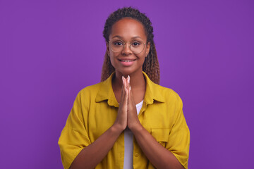 Young positive African American woman folds palms in front of chest making prayer gesture and looks...