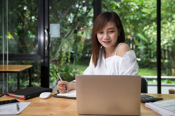 Satisfied woman writing in notebook and working using laptop at work. Woman with smile writing notes. Happy business woman looking at camera sitting inside office.