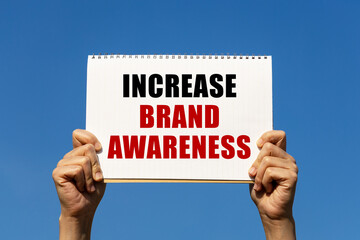Increase brand awareness text on notebook paper held by 2 hands with isolated blue sky background. This message can be used as business concept about increase brand awareness.